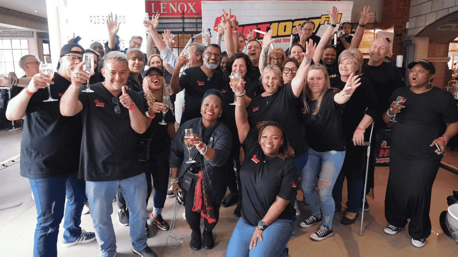 A large, diverse group of people in matching black shirts smiles and celebrates together indoors at Teddython 2025, raising glasses and posing enthusiastically for the camera.