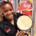 A smiling woman in a black shirt holds a small hand drum towards the camera, with colorful, music-themed posters from Olivedale Windmill displayed on the wall behind her.