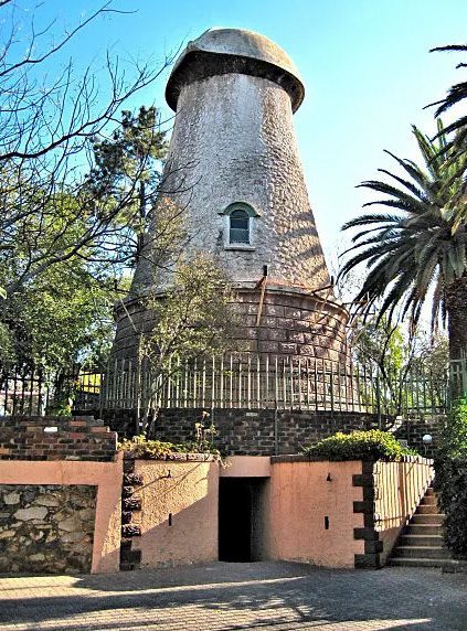 A historic windmill with a conical stone base and dome-shaped top stands behind a metal fence, offering unique character and charm—an inspiring symbol of homeownership—surrounded by trees, palm fronds, and an inviting stairway at the front.