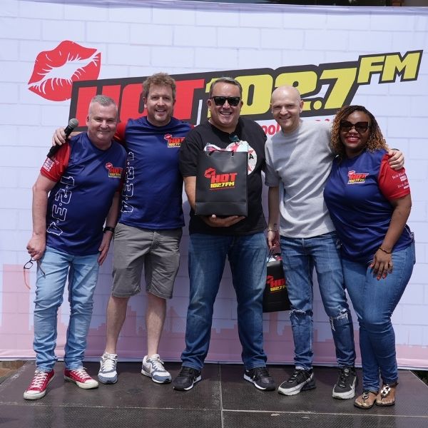 Five people stand smiling in front of a Hot 102.7 FM backdrop; one holds a branded gift bag. Casually dressed, they appear to be at a Radio Training Academy promotional event.