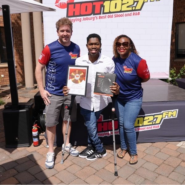 Three people smile outdoors in front of a HOT 102.7 FM banner. Two hold framed awards from the Radio Training Academy. The man on the left uses crutches, and all wear branded shirts except for the man in the middle, who is in a white shirt.