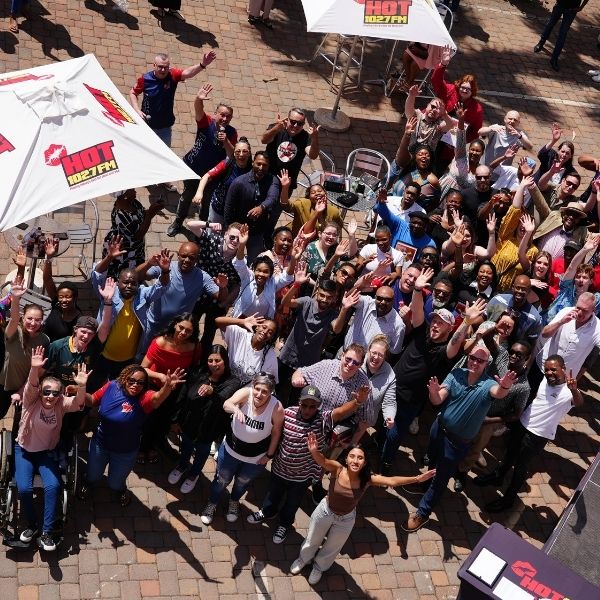 A large, diverse group of people stand outdoors on a brick patio, smiling and waving at the camera. Some tables and umbrellas with HOT 102.7FM logos are visible among the crowd at the Radio Training Academy event.