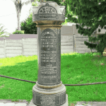 A tall cylindrical stone memorial stands on a lawn in the Magaliesberg area, engraved with multiple inscriptions and names. It is surrounded by a low metal railing, with trees, grass, and a fence in the background.