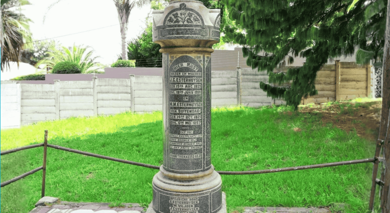 A tall cylindrical stone memorial stands on a lawn in the Magaliesberg area, engraved with multiple inscriptions and names. It is surrounded by a low metal railing, with trees, grass, and a fence in the background.
