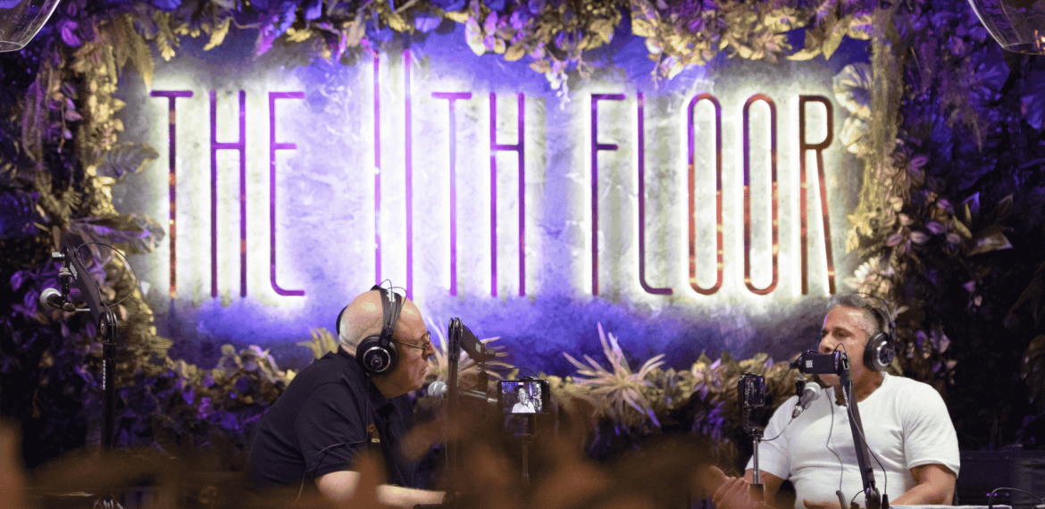 Two men wearing headphones and sitting at microphones discuss business opportunities in front of a sign that reads THE 11TH FLOOR, surrounded by decorative greenery and dramatic lighting.