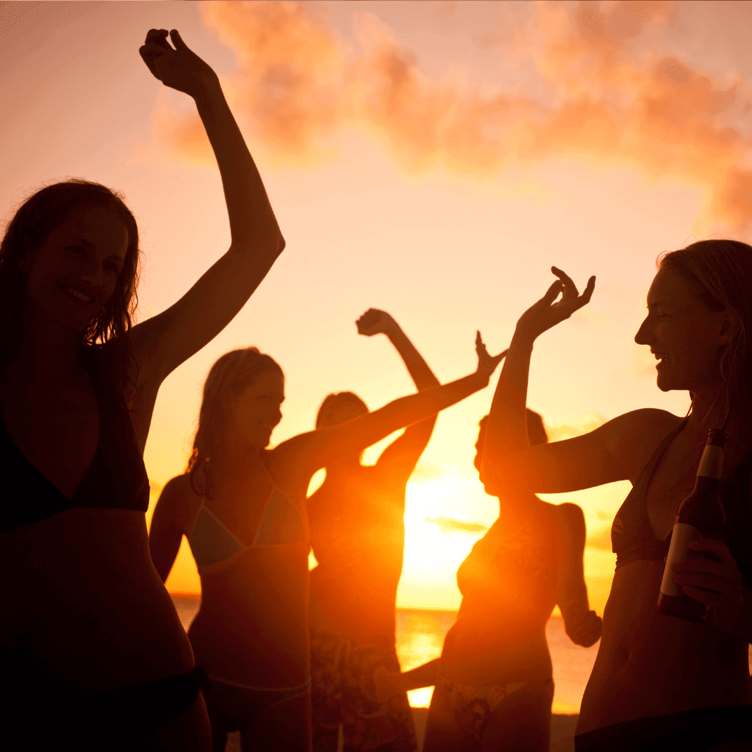 A group of people in swimsuits dance and celebrate on the beach at sunset, with warm orange light and silhouettes created by the sun low on the horizon. One person holds a bottle, toasting new business opportunities with friends.