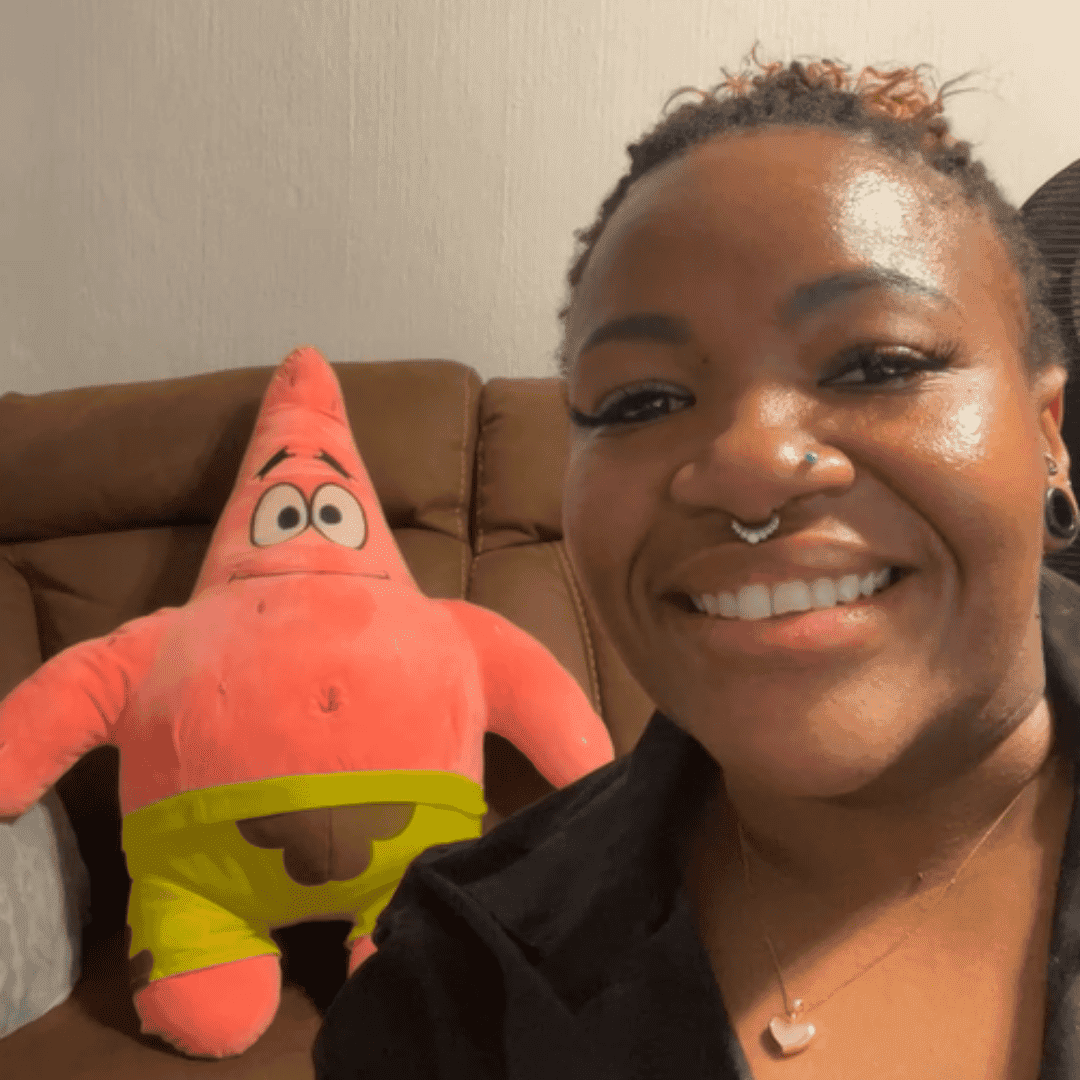 A smiling person with a nose ring and necklace takes a selfie on a couch with a large plush Patrick Star toy from SpongeBob SquarePants in the background, looking as cheerful as Sharon Corr at a concert.