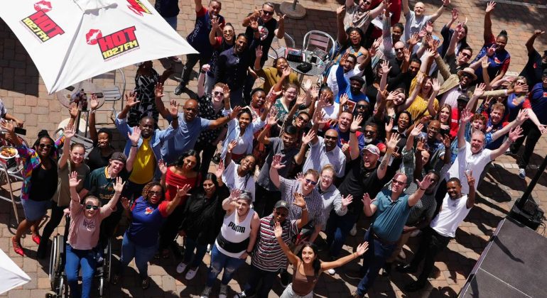 A large, diverse group of people outdoors on a patio, smiling and waving up at the camera. Some are seated, most are standing under umbrellas with HOT 1027 FM logos. It’s a bright, sunny day filled with excitement and new business opportunities.