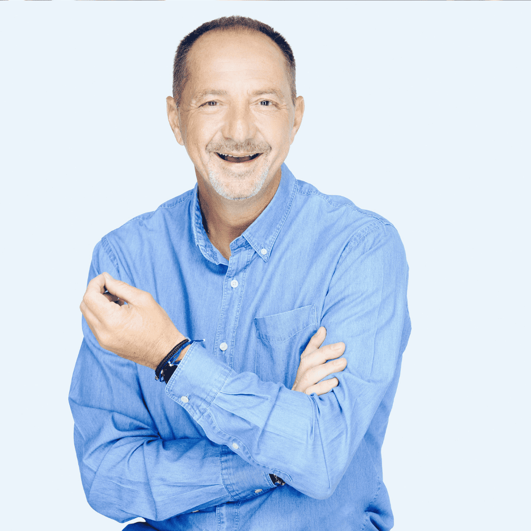 A middle-aged man with short hair and a mustache smiles in a light blue denim shirt, standing against a pale background. One arm crossed and the other raised, he looks as if he's chatting about plans for the weekends.
