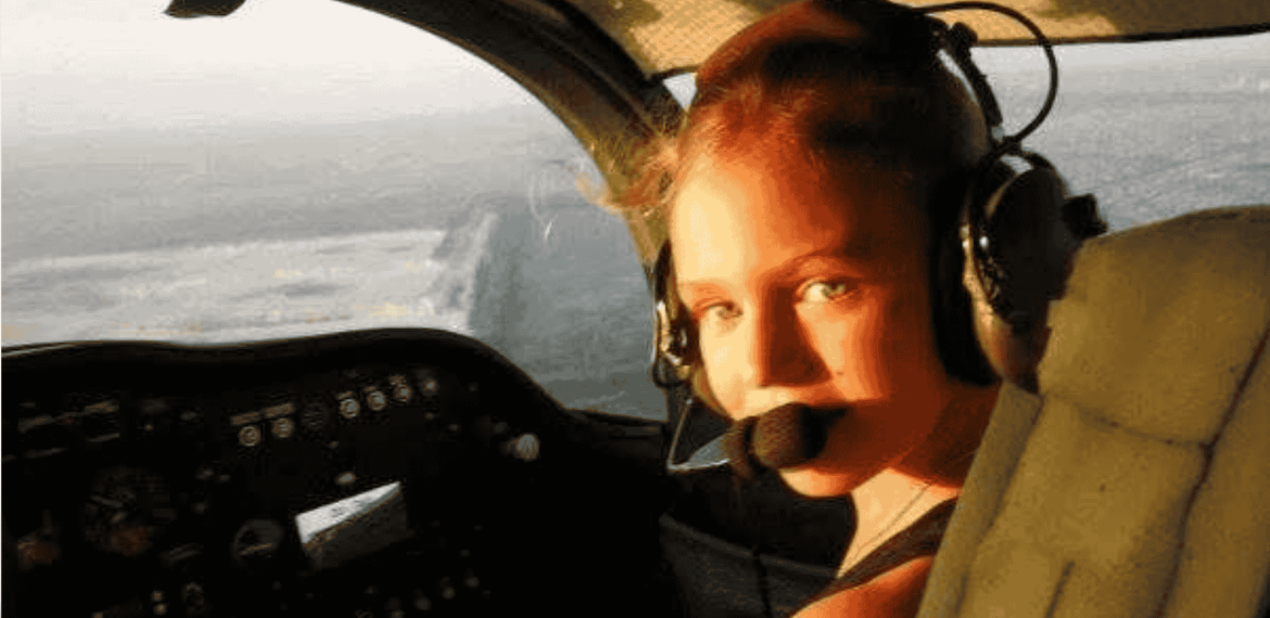 A young woman wearing a headset with a microphone sits in the cockpit of an airplane, looking back toward the camera as sunlight streams through the window, ready to explore new business opportunities.