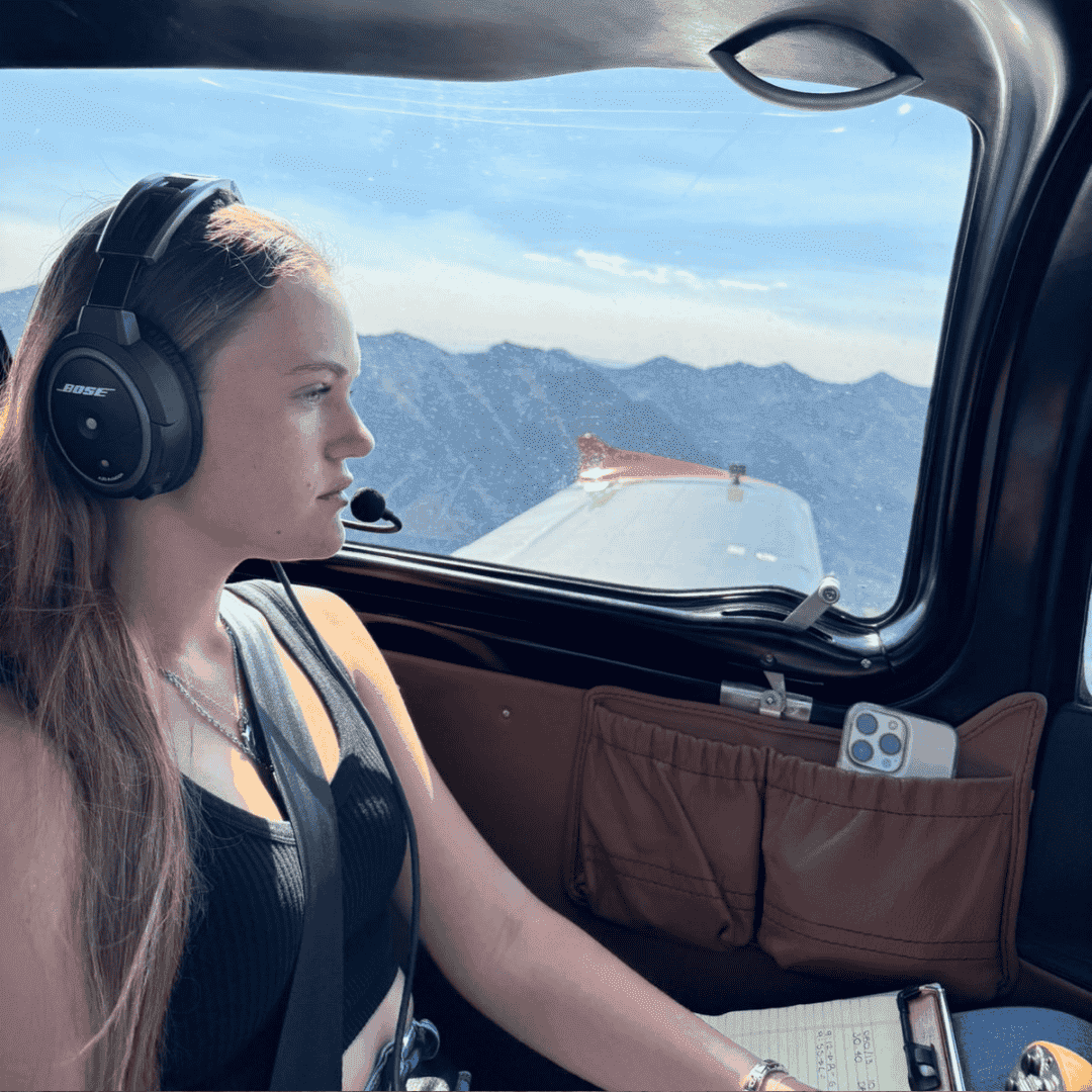 A woman wearing a headset sits in the cockpit of a small airplane, looking out the window at distant mountains under a clear sky. A notepad and pen rest on her lap, ready to jot down new business opportunities as she flies.