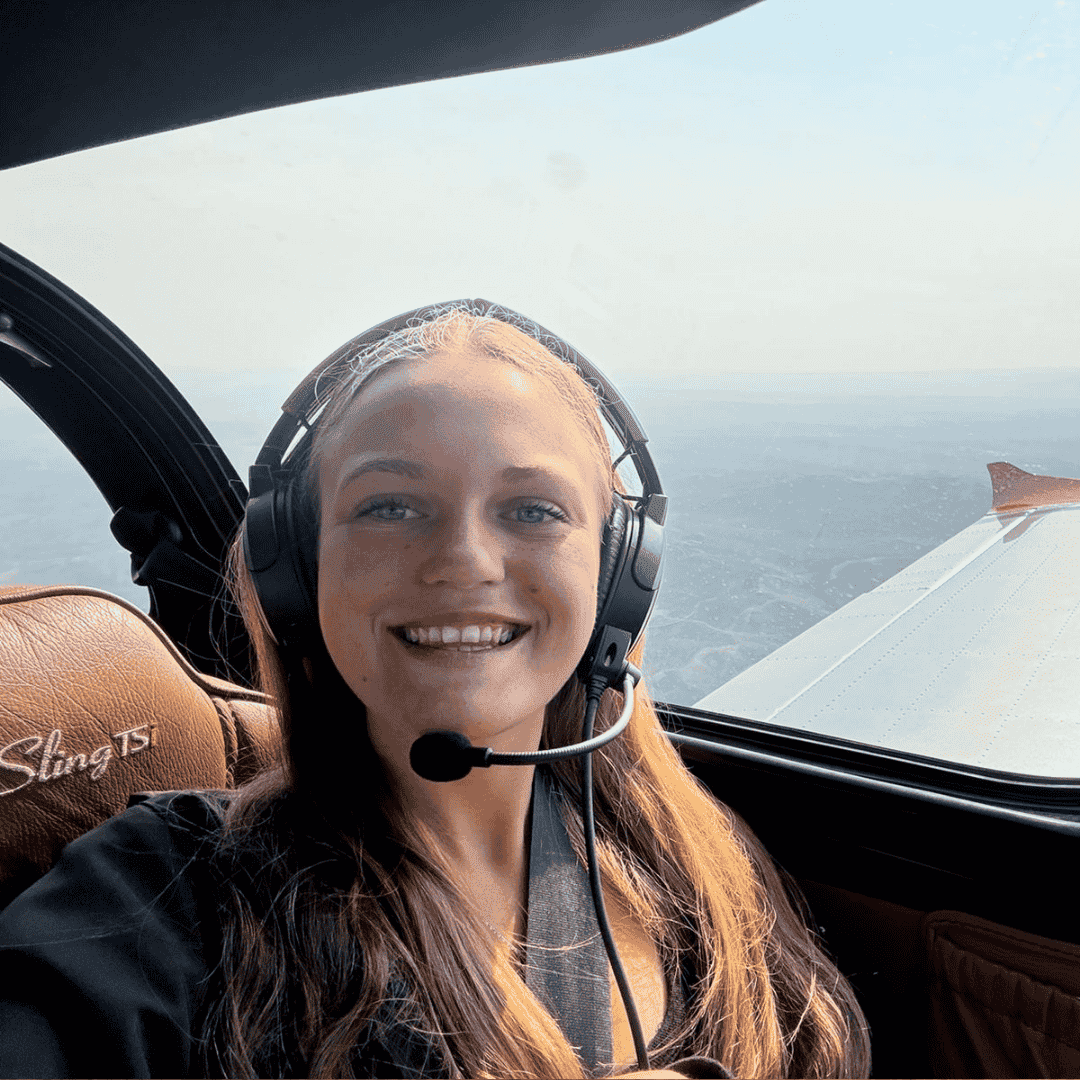 A smiling woman wearing a headset sits in the cockpit of a small plane, with the aircraft’s wing and blue sky visible through the window beside her—highlighting exciting business opportunities in aviation. The seat has Sling TS embroidered on it.