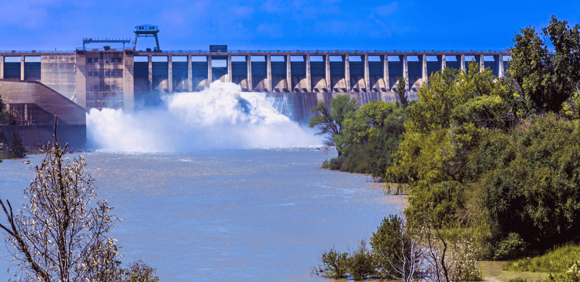A large dam releases water into a wide river, surrounded by green trees and bushes under the bright Magaliesberg sky with scattered clouds.