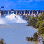 A large dam releases water into a wide river, surrounded by green trees and bushes under the bright Magaliesberg sky with scattered clouds.