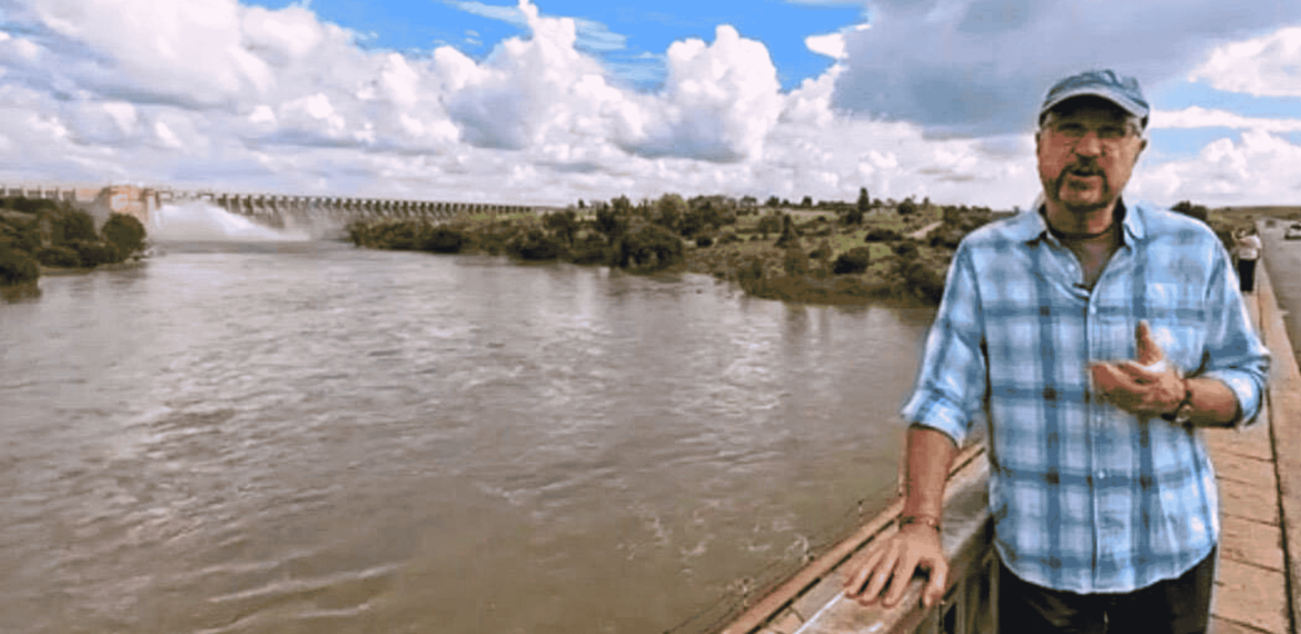 A man in a blue plaid shirt and cap stands on a bridge over a wide river near the Magaliesberg, with a dam and lush greenery in the background under a partly cloudy sky.
