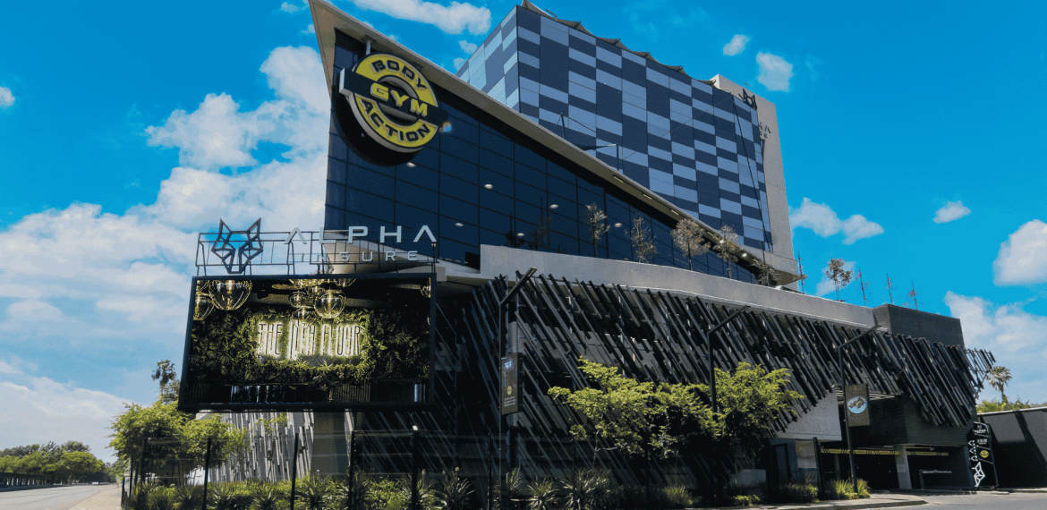 A modern, multi-story building with a geometric design and glass facade, featuring a large Body Action Gym sign and a digital billboard promoting new business opportunities, set under a bright blue sky with scattered clouds.