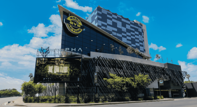 A modern, multi-story building with a geometric design and glass facade, featuring a large Body Action Gym sign and a digital billboard promoting new business opportunities, set under a bright blue sky with scattered clouds.