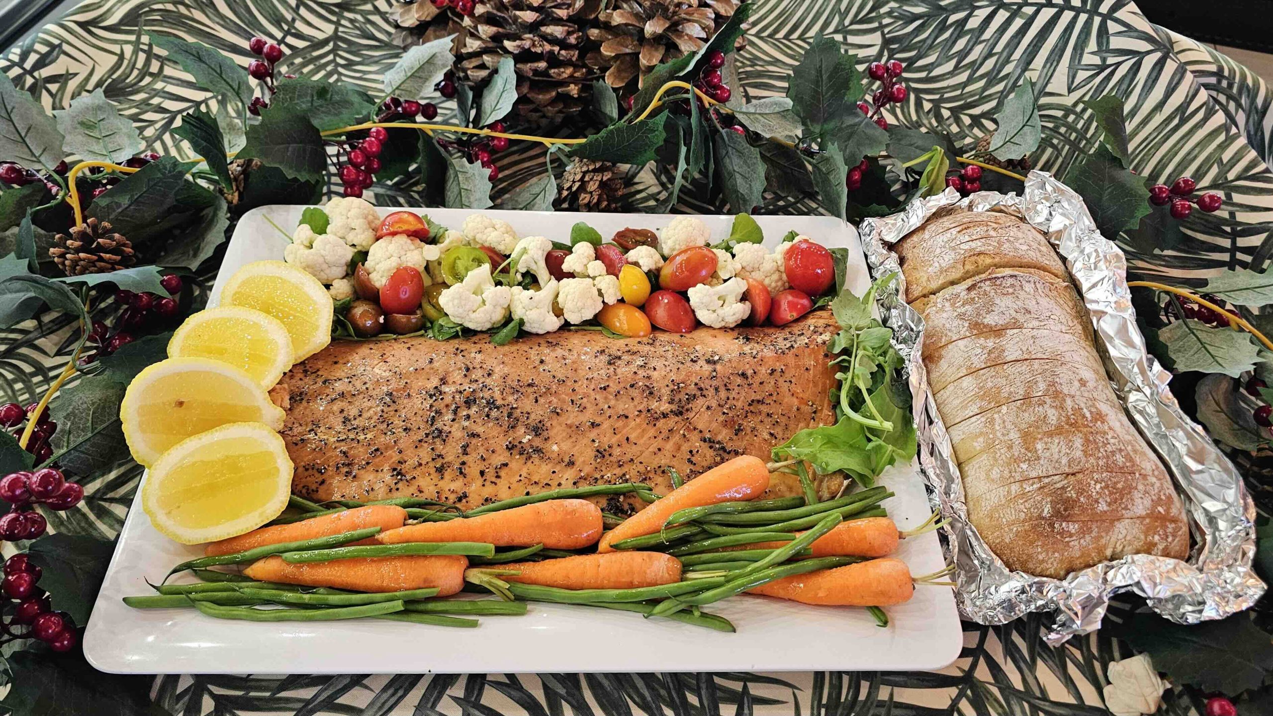 A foodie’s dream platter with seasoned baked salmon, lemon slices, baby carrots, green beans, cherry tomatoes, and cauliflower, paired with a foil-wrapped baguette and garnished with greenery, berries, and pinecones.