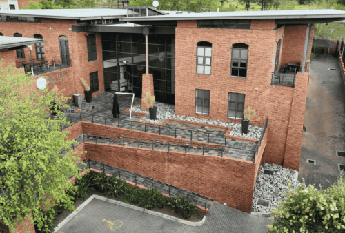 A modern red-brick building with large windows, outdoor potted plants, and a ramp along the front, surrounded by greenery and a paved driveway—ideal for those seeking new business opportunities.