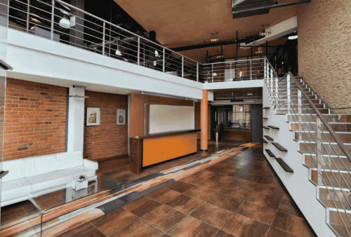 Modern office lobby with brown tile floors, orange and white reception desk, white leather seating, and a staircase with metal railings leading to a mezzanine level—an inviting space ideal for new business opportunities.