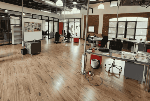 A spacious, modern office with wooden floors, scattered desks and chairs, large windows, exposed brick walls, and glass-walled meeting rooms. The bright, organized space is ideal for fostering creativity and exploring new business opportunities.