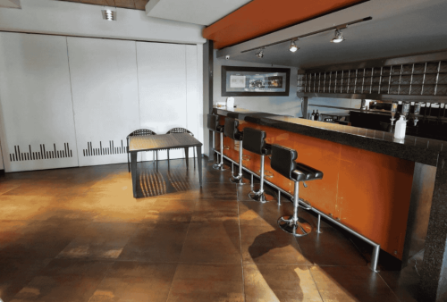Modern kitchen with an orange and black bar counter, four black bar stools, a small black dining table with two chairs, brown tile flooring, wall art, and a row of glass shelves behind the counter—ideal for exploring business opportunities.