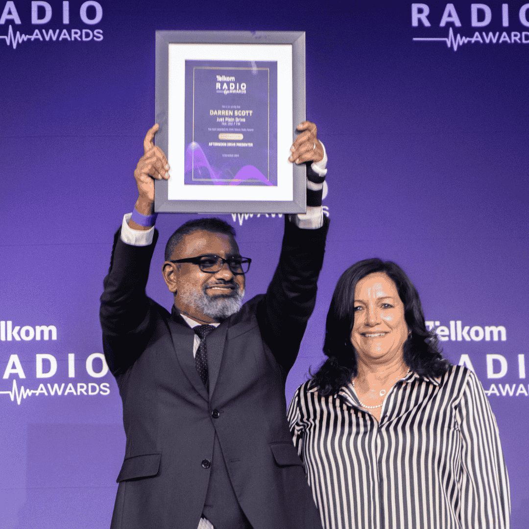A man in a suit and tie holds up a framed award and smiles beside a woman in a striped blouse, standing in front of a purple Telkom Radio Awards backdrop, celebrating their success near the Vaal Dam.