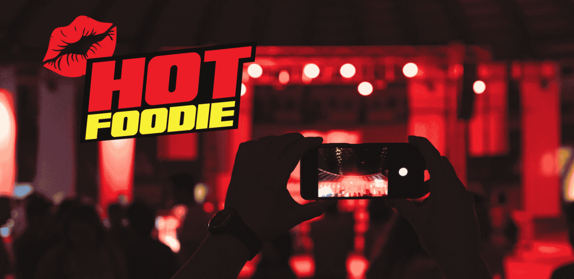A person holds up a smartphone to take a photo at a red-lit event, with the words HOT FOODIE and a red lips graphic displayed prominently on the left side.