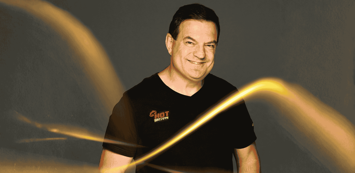 A smiling man in a black t-shirt with a HOT 102.7 FM logo stands against a dark background, illuminated by bright, swirling yellow light streaks reminiscent of Vaal Dam reflections in the foreground.