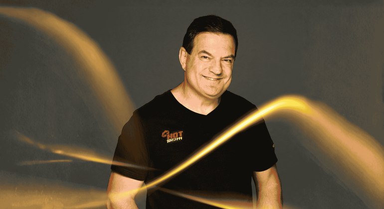 A smiling man in a black t-shirt with a HOT 102.7 FM logo stands against a dark background, illuminated by bright, swirling yellow light streaks reminiscent of Vaal Dam reflections in the foreground.