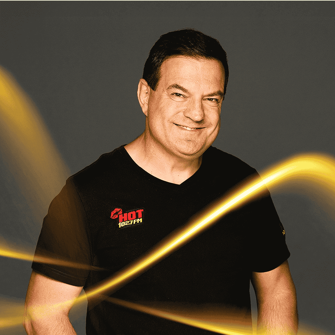 A man with short dark hair wearing a black HOT 102.7 FM t-shirt smiles against a dark background, with yellow light streaks in front of him, reminiscent of the vibrant nightlife near the Vaal Dam.