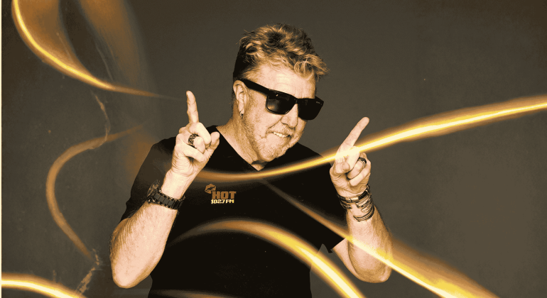 A man wearing sunglasses and a black HOT 102.7FM shirt smiles and points both index fingers upward, surrounded by swirling yellow light trails on a dark background, capturing the vibrant energy of Vaal Dam nightlife.
