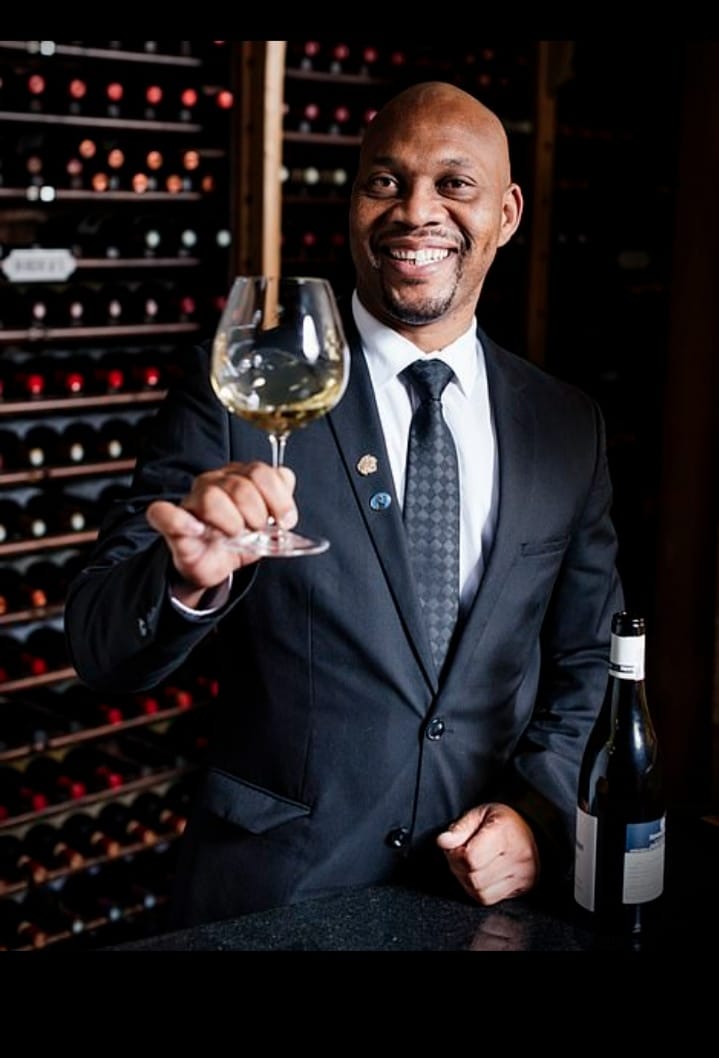 A smiling foodie in a suit holds up a glass of white wine in front of shelves filled with wine bottles, with an open wine bottle on the counter beside him.