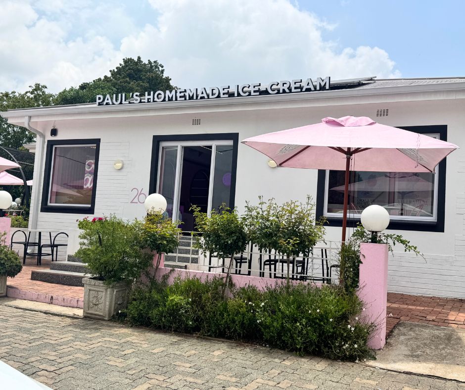 A white building with “PAUL’S HOMEMADE ICE CREAM” sign, pink umbrellas shading dog friendly outdoor tables, and green bushes lining the entrance. The setting appears bright and welcoming under a partly cloudy sky.