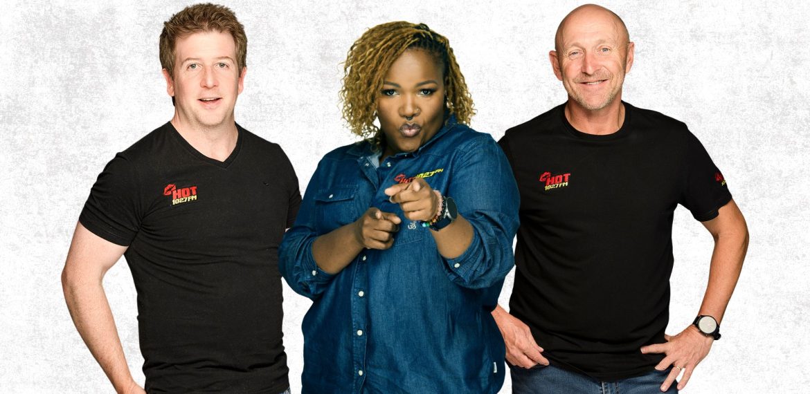 Three people stand side by side against a white background. The two men wear black SHOT FACTORY shirts, while the woman in a blue denim shirt points and looks toward the camera, reflecting a dynamic cross-border creative team.