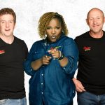 Three people stand side by side against a white background. The two men wear black SHOT FACTORY shirts, while the woman in a blue denim shirt points and looks toward the camera, reflecting a dynamic cross-border creative team.