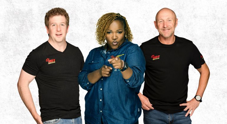 Three people stand side by side against a white background. The two men wear black SHOT FACTORY shirts, while the woman in a blue denim shirt points and looks toward the camera, reflecting a dynamic cross-border creative team.