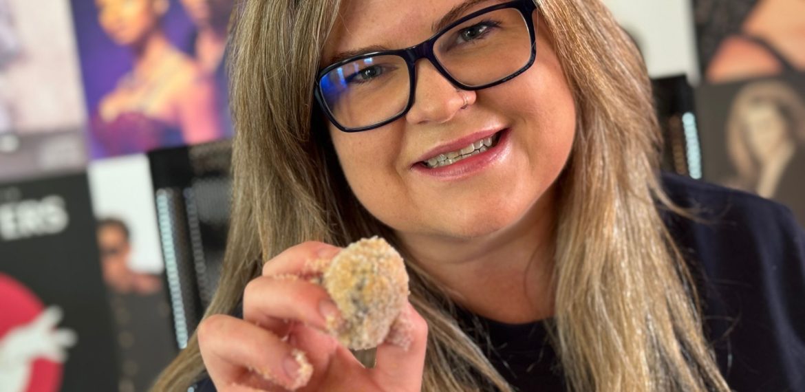 A woman with long blonde hair and glasses smiles at the camera, holding up a small breaded Matric snack. Blurred music posters are visible in the background.