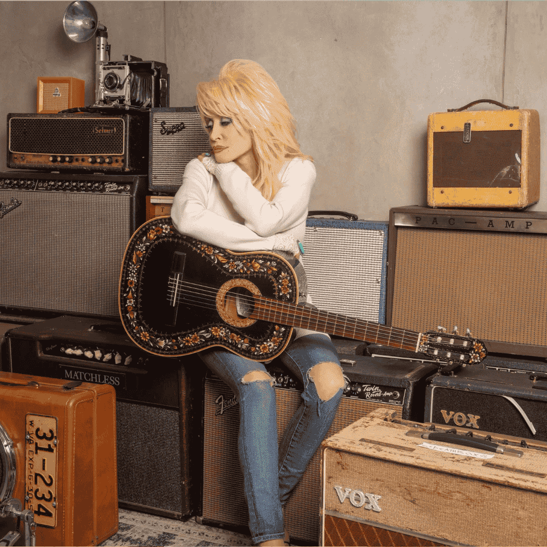 A person with long blonde hair sits on an amplifier, holding a black acoustic guitar adorned with floral designs, surrounded by vintage amplifiers and speakers in a cozy, music-themed room—a perfect matric celebration setting.