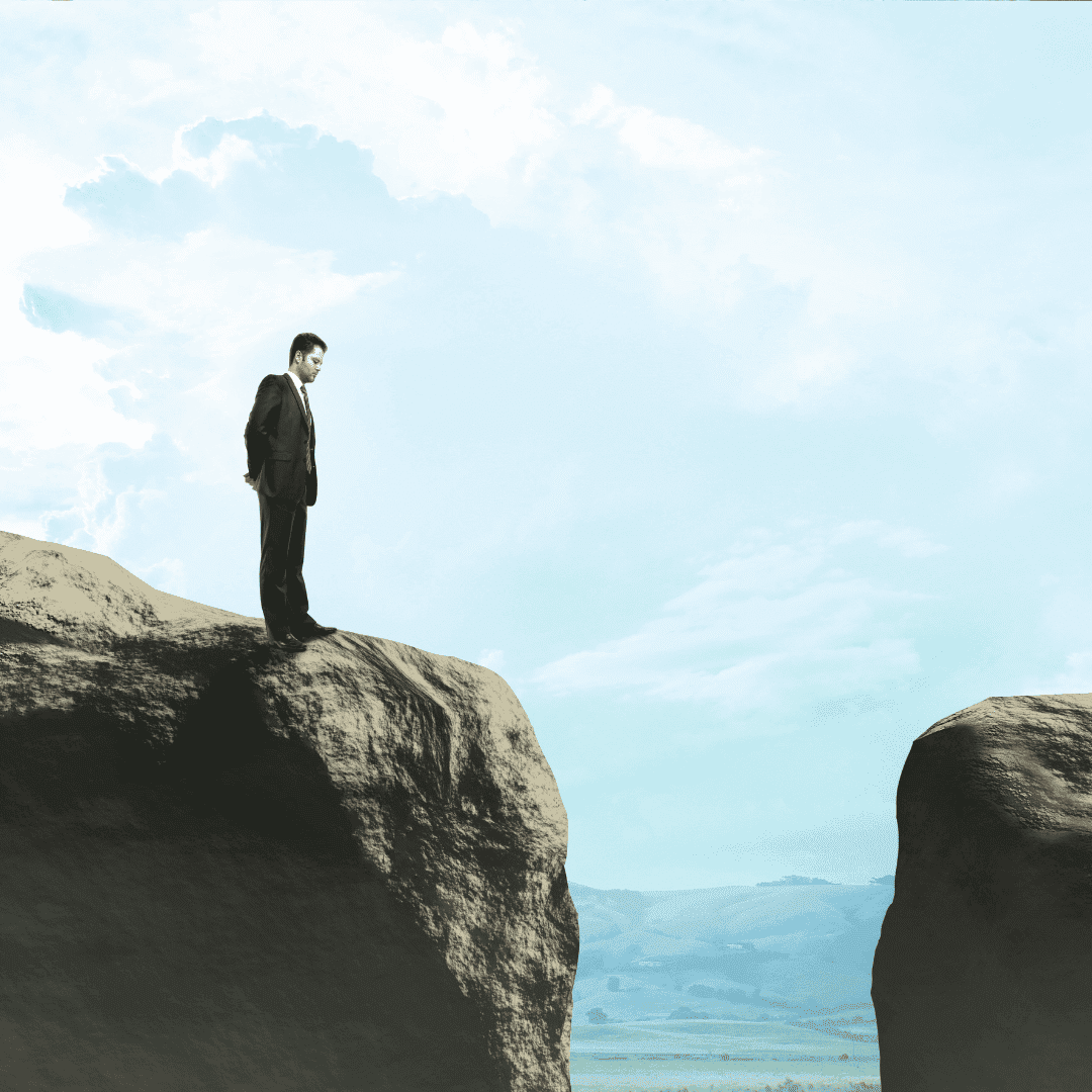 A man in a suit stands on the edge of a rocky cliff, looking down at a wide gap separating him from new business opportunities on another cliff under a bright, cloudy sky.
