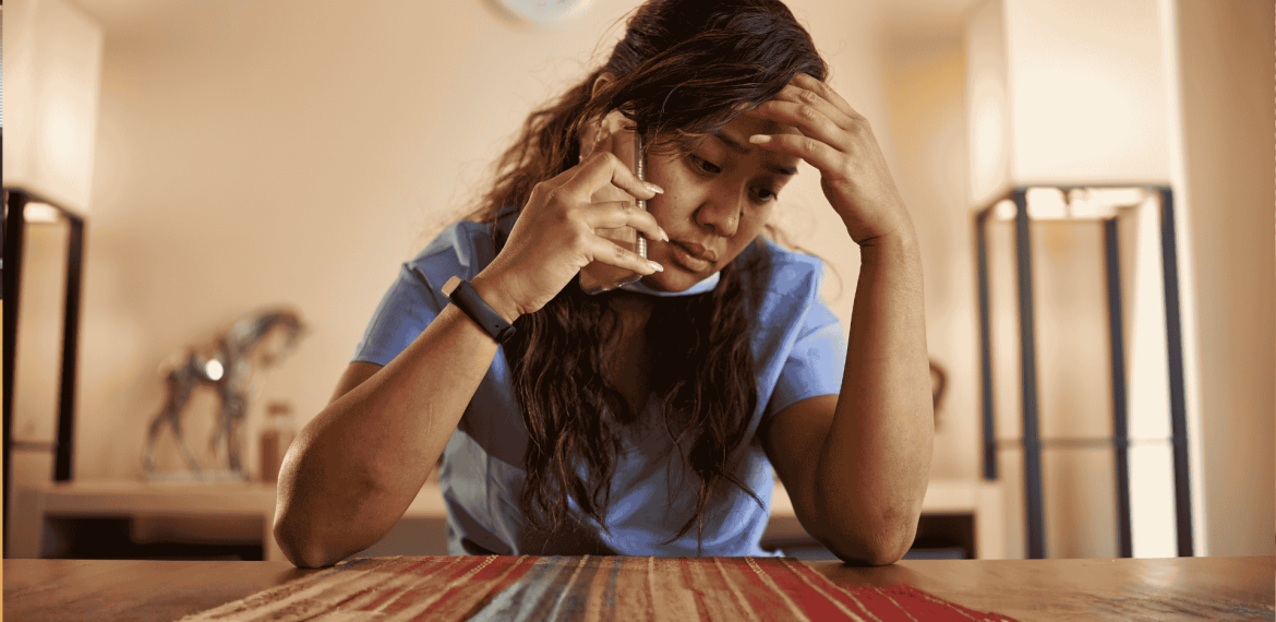A woman in scrubs sits at a table, looking stressed while talking on the phone about business opportunities, her hand resting on her forehead.