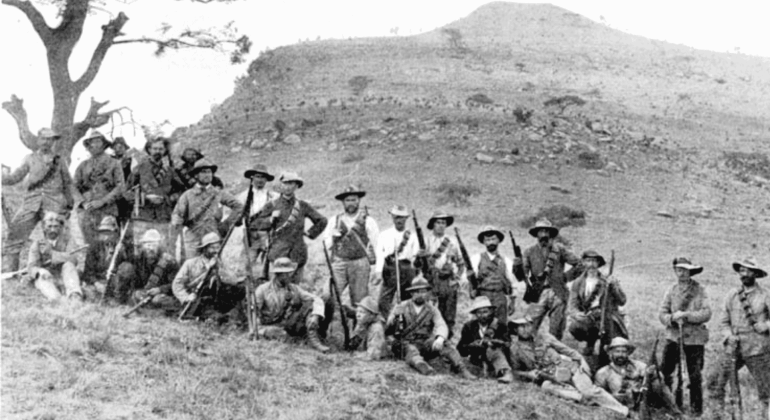 A group of men in hats and old-fashioned clothing pose with rifles on a grassy hillside near Yeoville; a lone tree stands to the left and a rugged hill rises in the background. The photo appears to be historical.