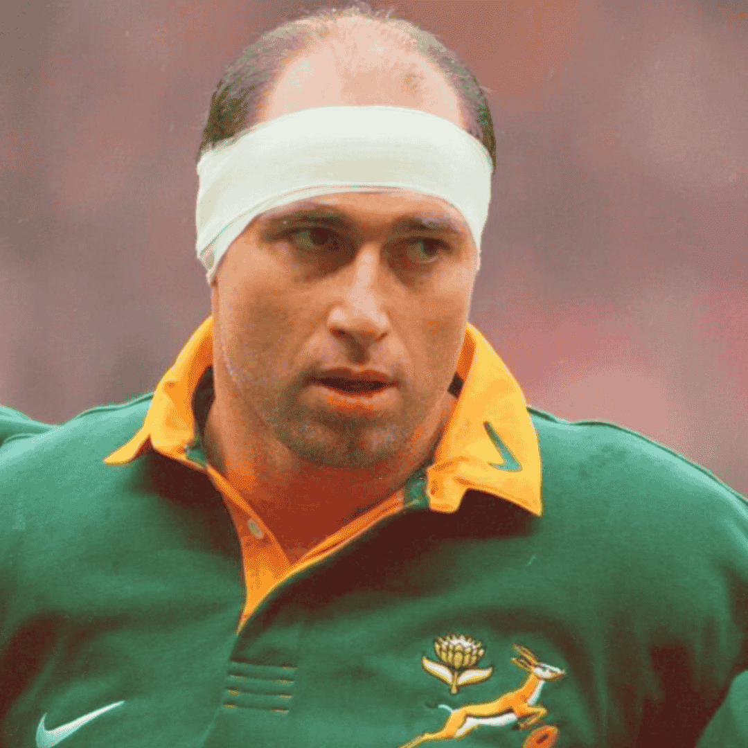 A rugby player in a green and yellow jersey with a Springbok logo stands with a focused expression, wearing a white bandage around his head, ready for intense cross-border competition.