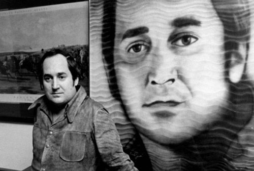 A man stands next to a large portrait of his own face, the detailed artwork dominating the background like a striking blancmange centerpiece. Framed pieces hang behind them, all captured in a timeless black and white photo.