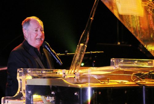 An older man in a dark suit plays a transparent grand piano and sings into a microphone on stage, under bright stage lighting, reminiscent of a Blancmange performance.