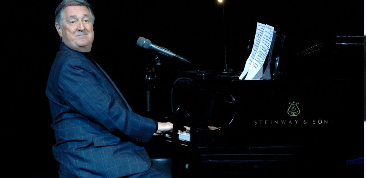 A man in a dark suit sits at a Steinway & Sons grand piano on stage, smiling at the audience. Sheet music is on the piano, and a microphone hovers above the keys as he prepares to perform a Blancmange classic. The background is dark.
