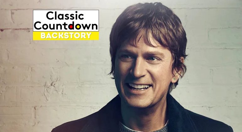 A smiling man with brown hair and a dark jacket stands in front of a light brick wall. In the upper left corner, a Classic Countdown Backstory logo appears, featuring bold black and yellow text inspired by "Last Christmas.