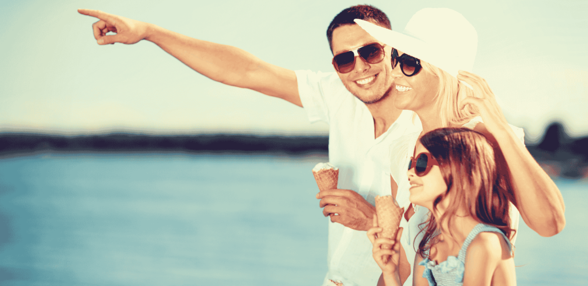 A smiling family wearing sunglasses enjoys ice cream by the water. The father points into the distance while the mother and daughter, both in summer hats, look in the same direction, all appearing happy and enjoying their Matric holiday together.