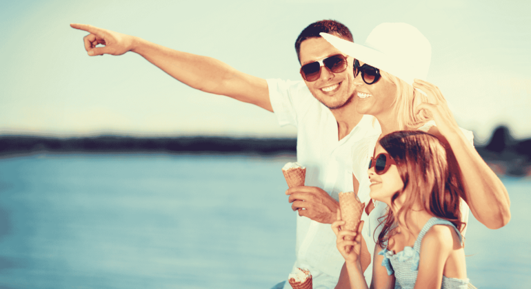 A smiling family wearing sunglasses enjoys ice cream by the water. The father points into the distance while the mother and daughter, both in summer hats, look in the same direction, all appearing happy and enjoying their Matric holiday together.