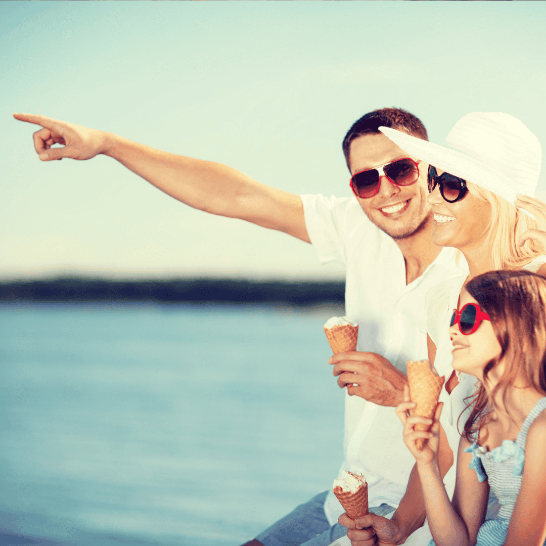 A smiling family of three in sunglasses enjoys ice cream cones by the water. The man, who just celebrated his matric results, points into the distance while the woman and girl stand beside him, all wearing summer clothes and looking happy.